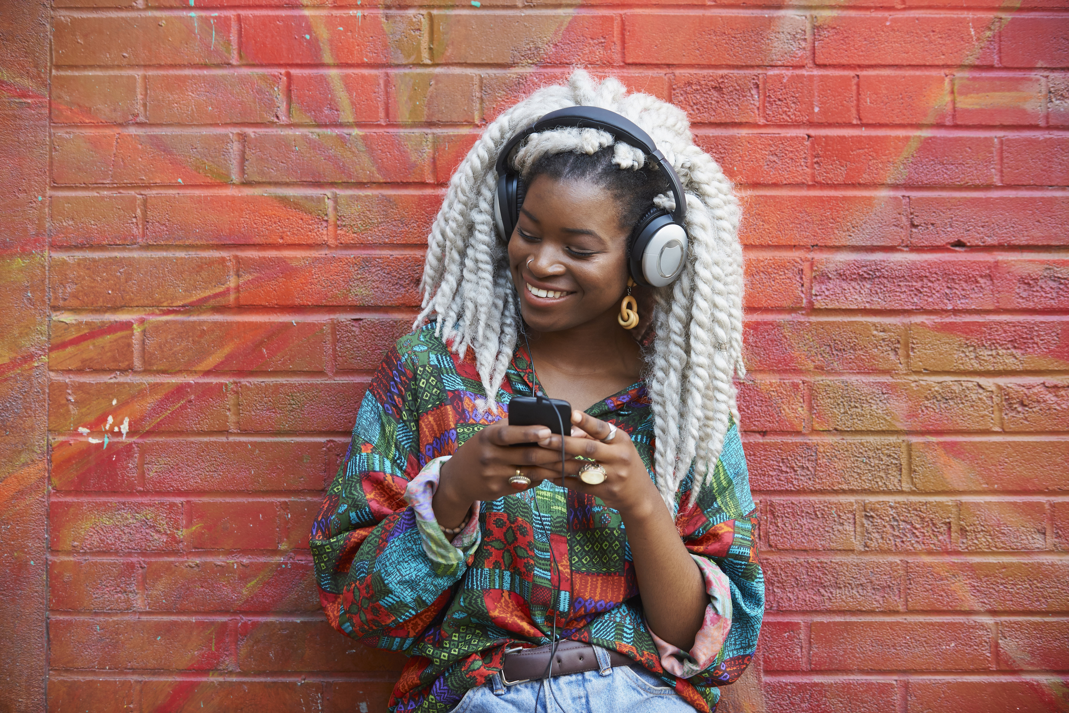 Black woman leaning on brick wall listening to cell phone with headphones