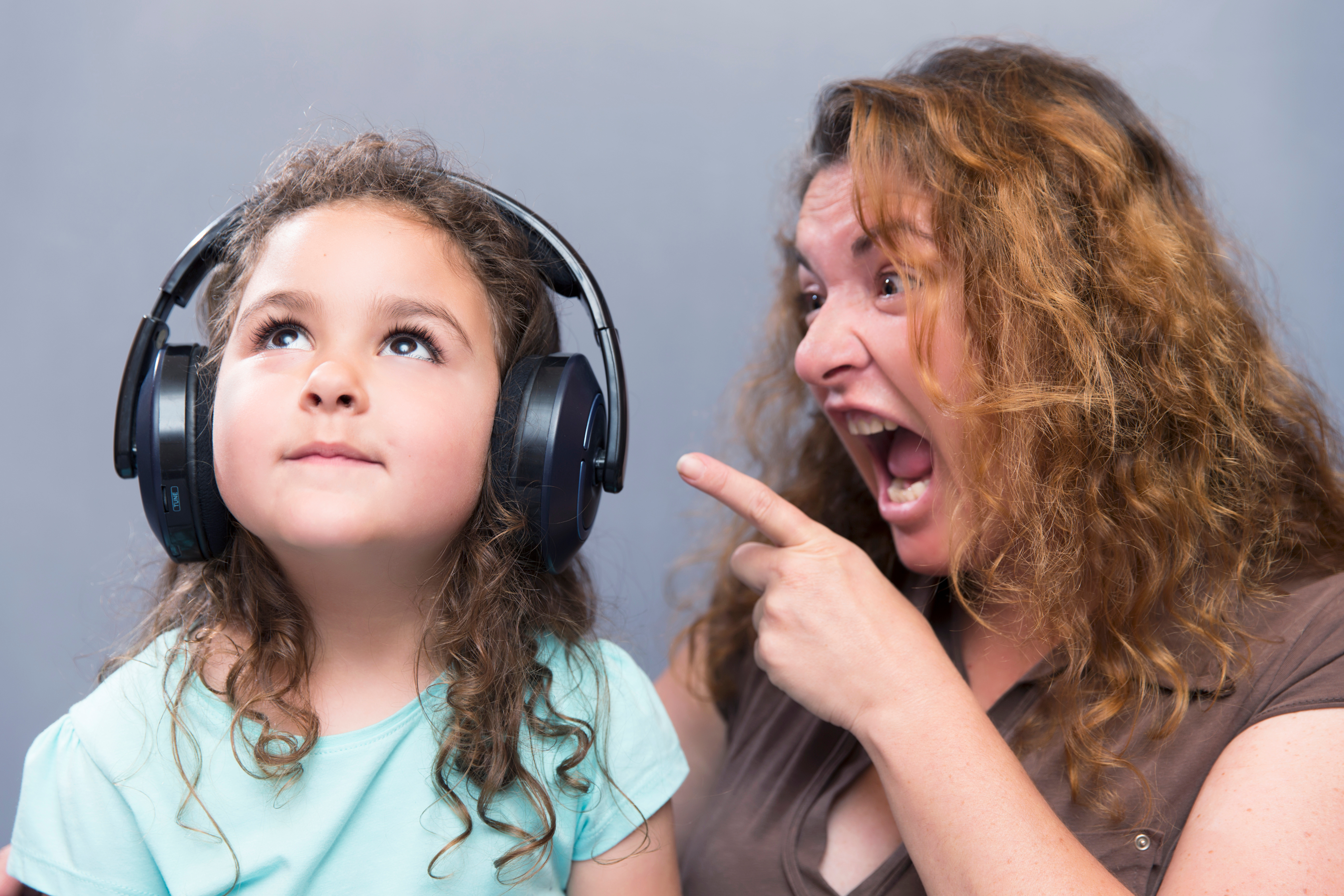 Angry Mother Shouting While Cute Girl Listening To Headphones Against Gray Background