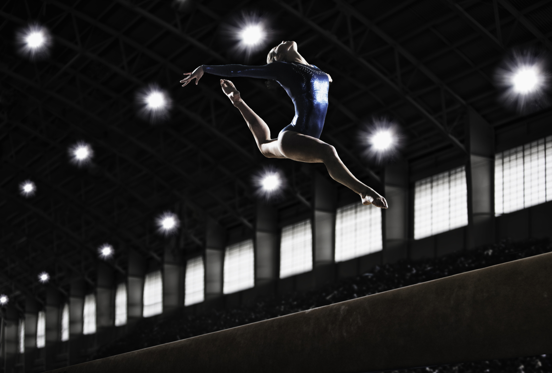 A female gymnast, a young woman performing a floor routine, in mid air with legs and arms outstretched and back bent.