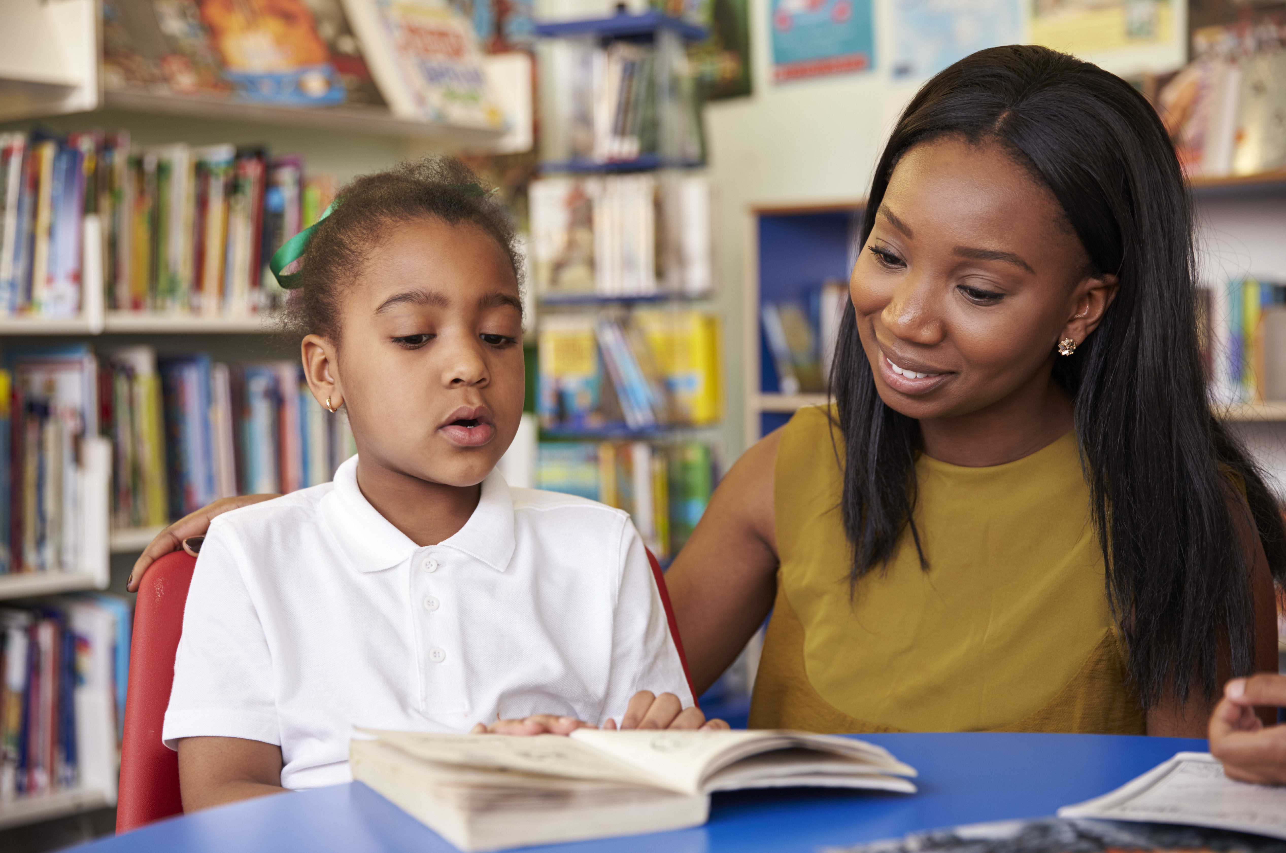 Elementary School Pupil Reading In Library With Teacher