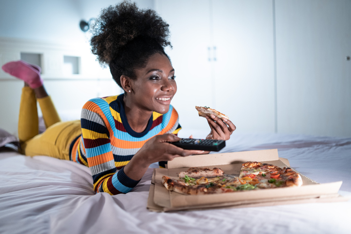 Afro woman spending lonely evening in bed with pizza and watching tv