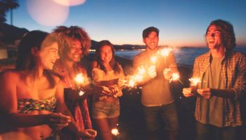 Group of friends playing with sparklers and fireworks on the beach at sunset.