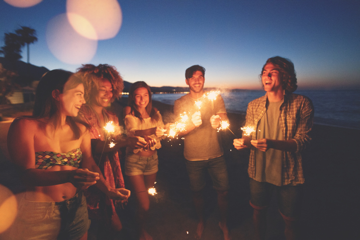 Group of friends playing with sparklers and fireworks on the beach at sunset.