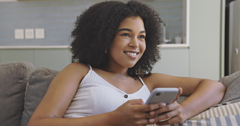 Beautiful young black woman using her phone while sitting on the couch in her living room at home. Happy female with afro thinking what to reply while texting or browsing through social media posts