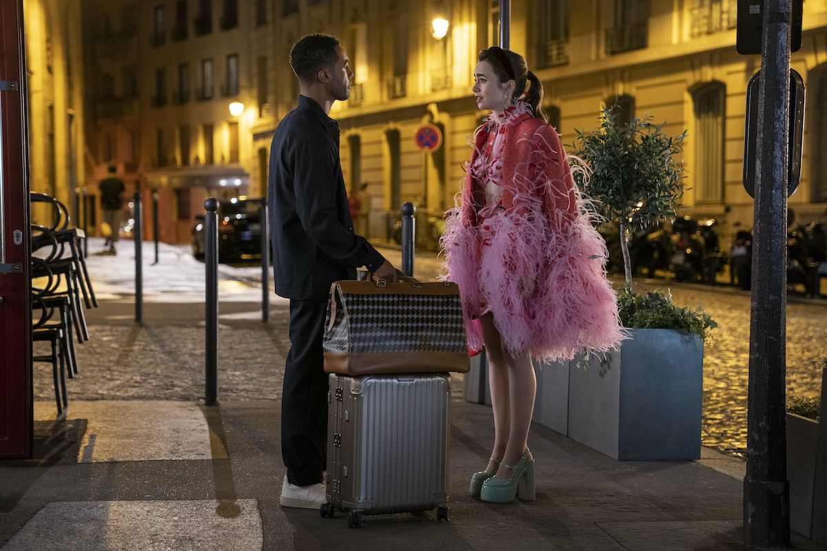 Emily In Paris production still from Season 3 showing Lucien Laviscount as Alfie having a serious chat with Lily Collins as Emily while holding his luggage
