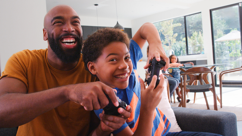 African American Father And Son Sitting On Sofa At Home Playing Video Game Together