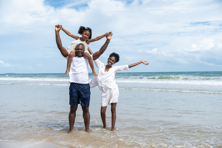 Happy black family father mother daughter smiling togetherness on beach with ocean waves and blue sky. Parents with child enjoying fun summer holiday